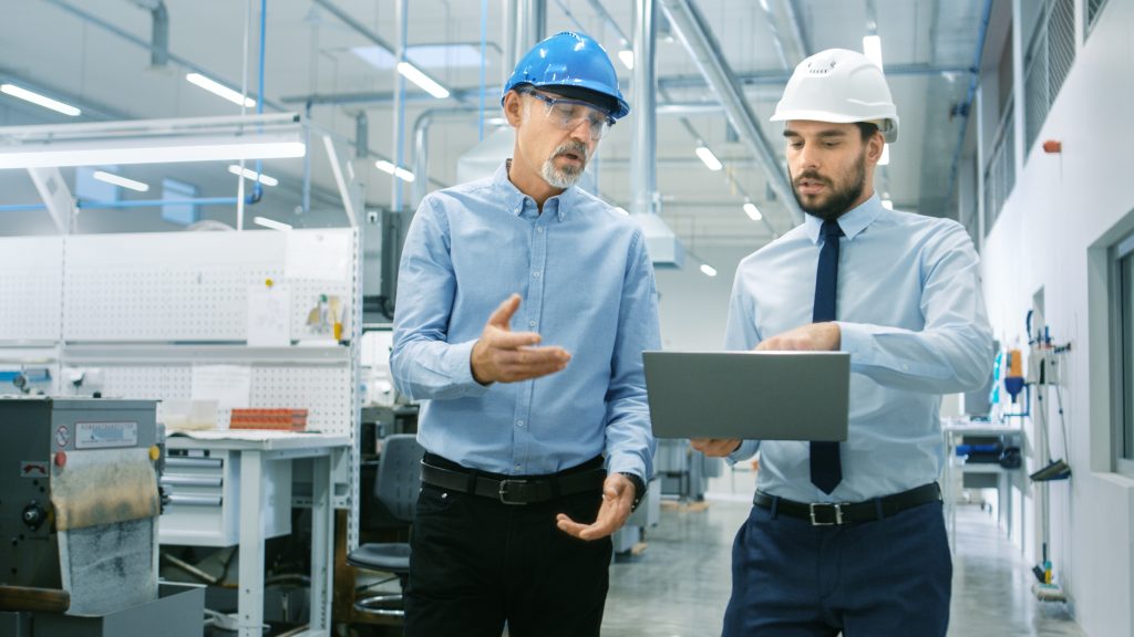 Two men in hard hats walk a manufacturing facility while looking at and holding a laptop.